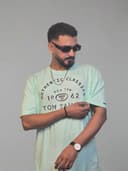 Man wearing a light blue t-shirt with text, sunglasses, and a watch, lying on a white surface with green leaves.