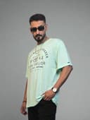 Man in light green shirt and black pants standing next to a plant indoors.