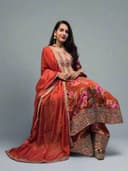 Woman in a traditional outfit with floral patterns, standing indoors with plants around.