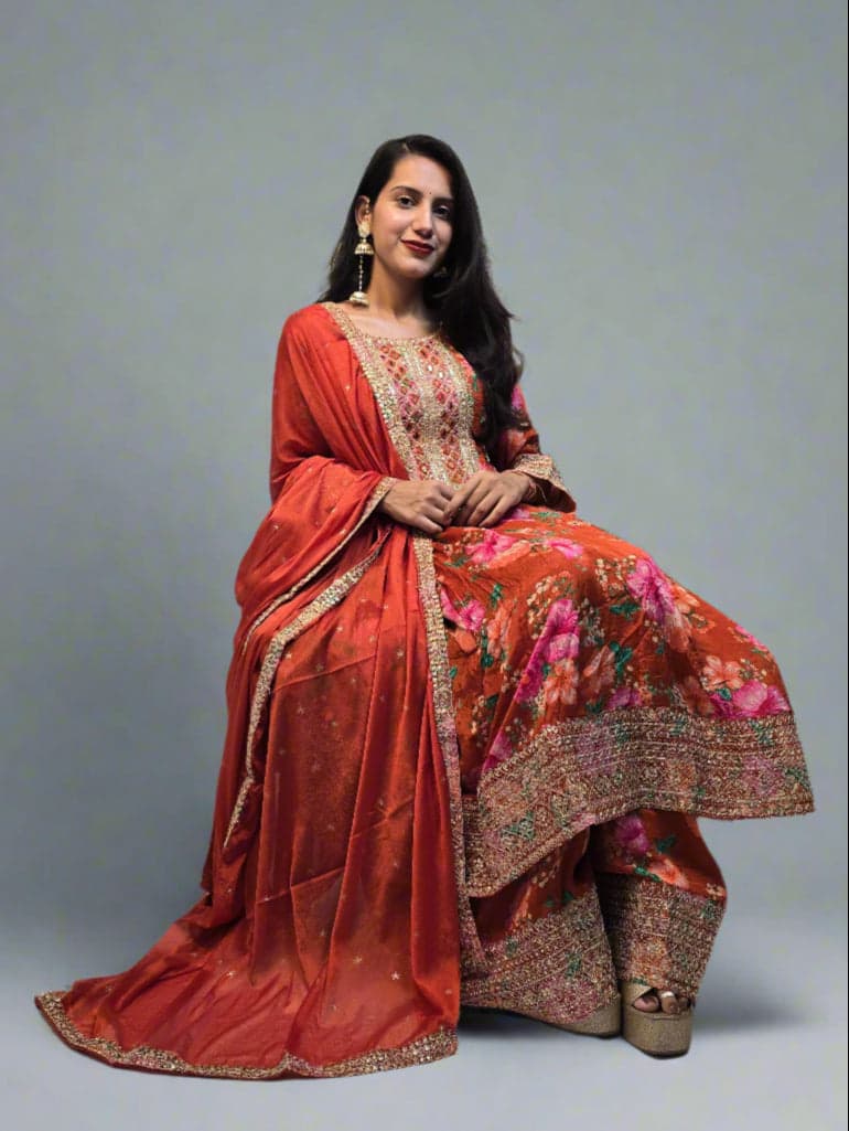 Woman in a traditional outfit with floral patterns, standing indoors with plants around.