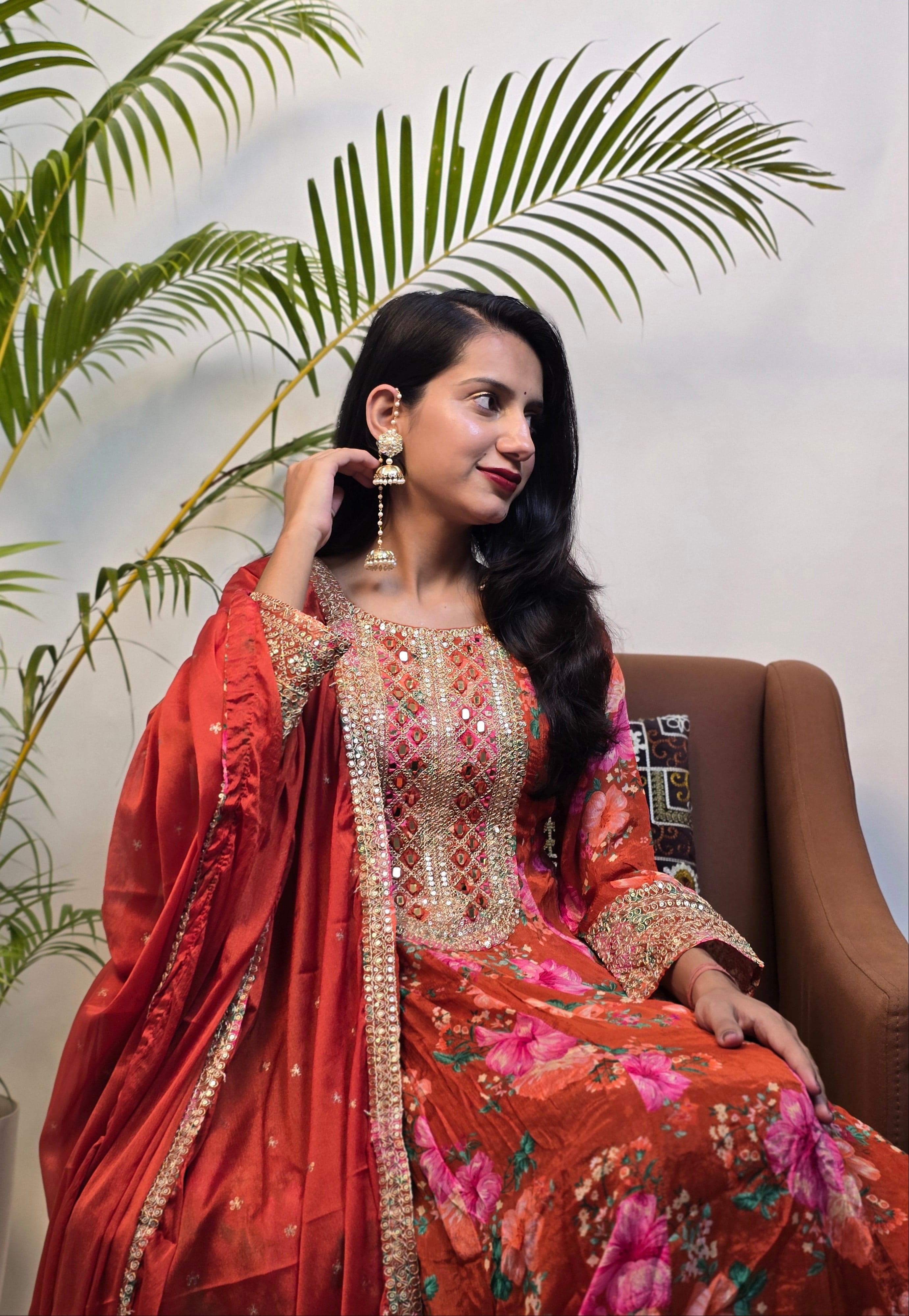Woman in traditional red and gold outfit with floral patterns, standing against a white background with green leaves.