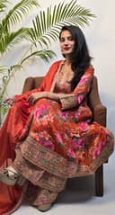 Woman in traditional outfit with floral patterns sitting on a white surface with green leaves.