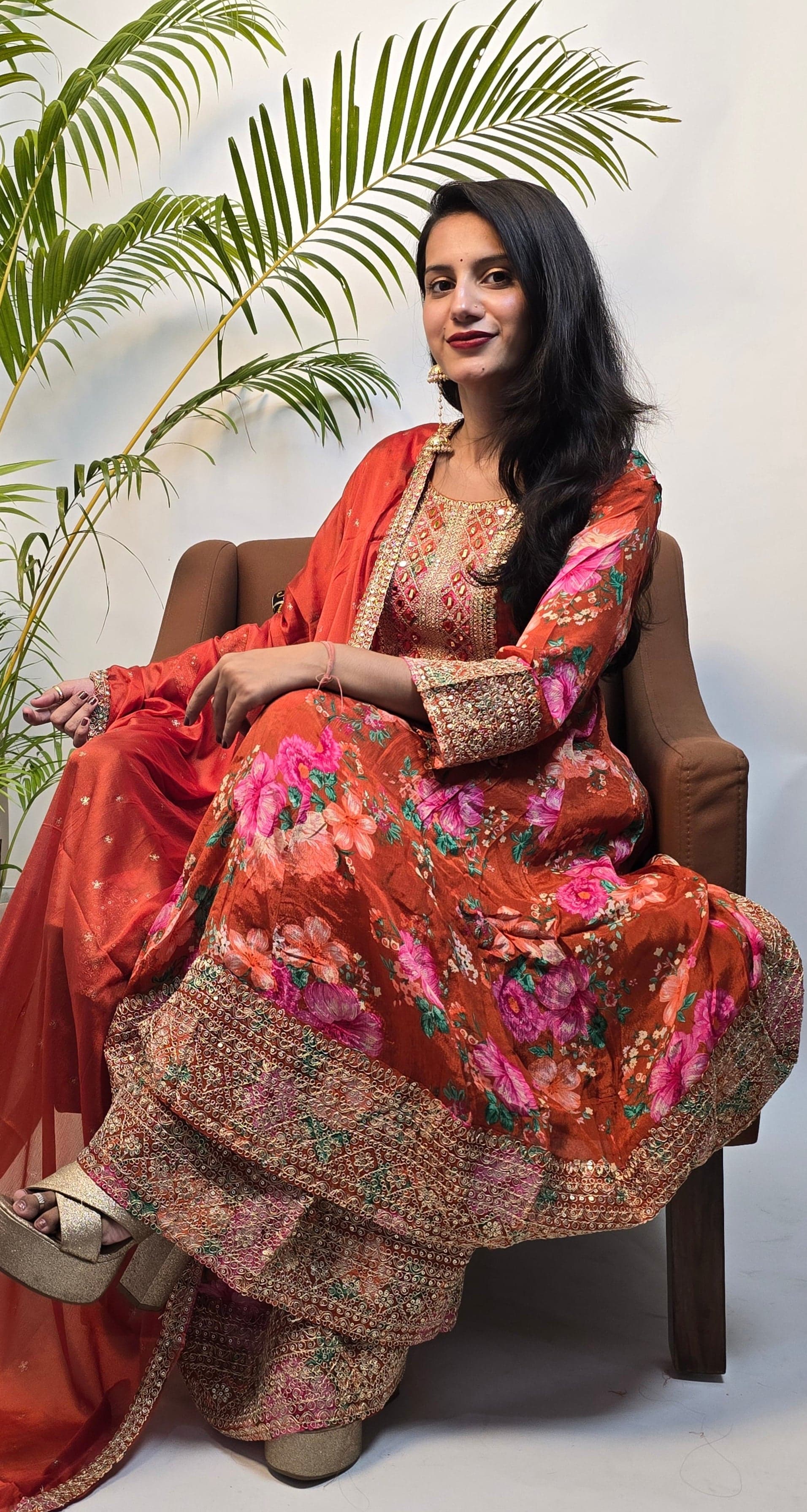 Woman in traditional outfit with floral patterns sitting on a white surface with green leaves.