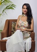 Woman sitting on a brown chair with a white garment draped over her, against a plain background.
