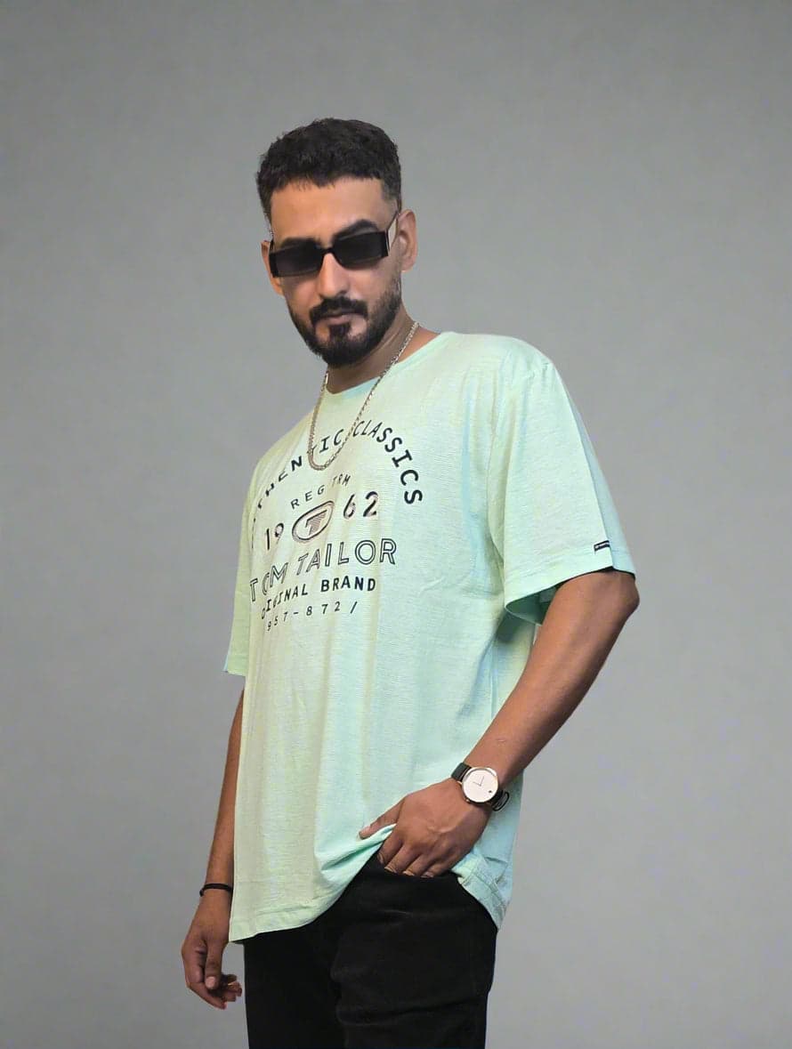 Man in light green shirt and black pants standing next to a plant indoors.