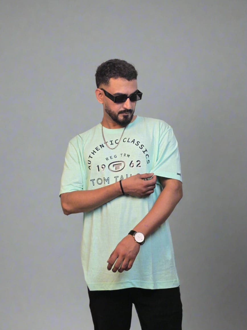 Man wearing a light blue t-shirt with text, standing against a neutral background with greenery.